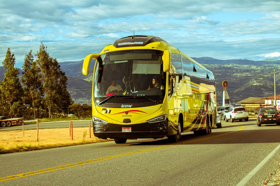 A bright yellow bus travels on a scenic mountain road under a blue sky, surrounded by cars and nature.